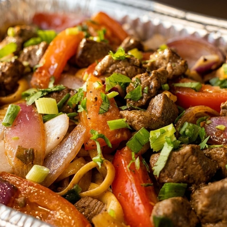 Close-up of Lomo Saltado with beef, red onions, tomatoes, and noodles in a foil container.